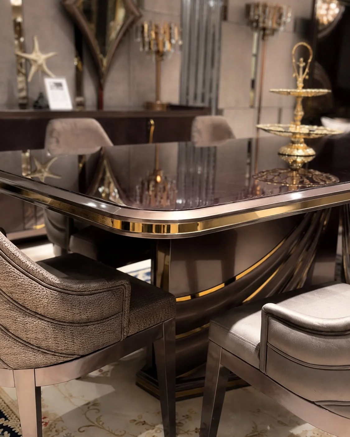 Close-up of the Shawl glossy dark dining table with rounded corners, gold-tone trim, sweeping sculptural pedestal and taupe chairs