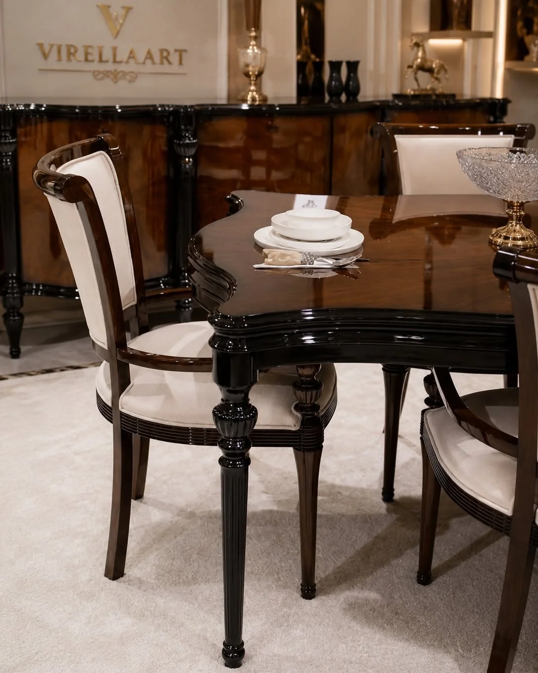 Close-up of the Valdera dining table corner with a glossy walnut-toned top, black carved edge, turned leg and cream upholstered chair