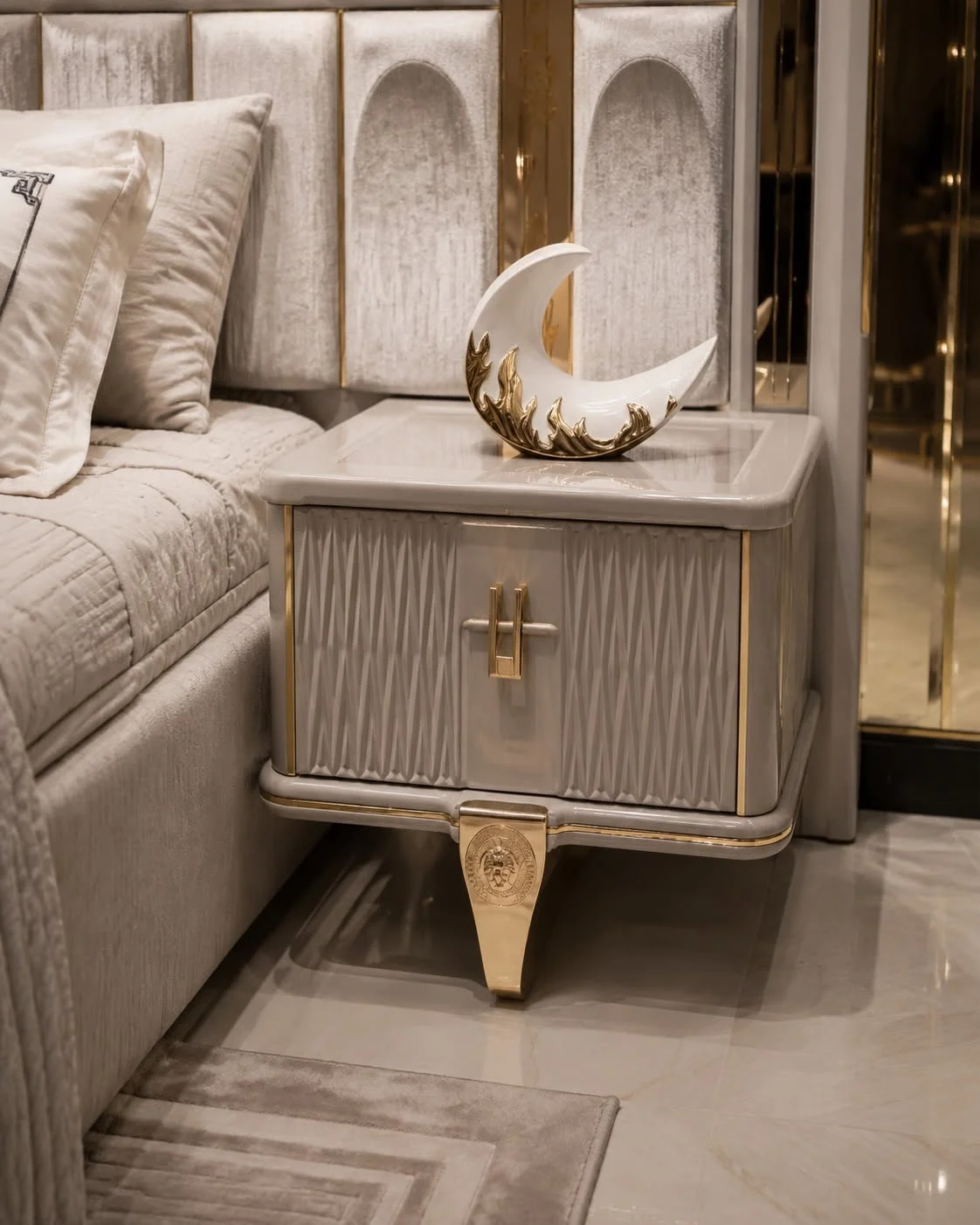 Close-up of the Verona cream nightstand with textured drawer front, gold-tone handle and tapered decorative leg beside the upholstered bed
